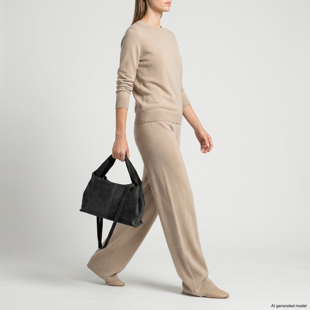 A person in a beige sweater and matching wide-leg pants walks while carrying a large black suede handbag, showcasing Italian craftsmanship. The background is plain white.