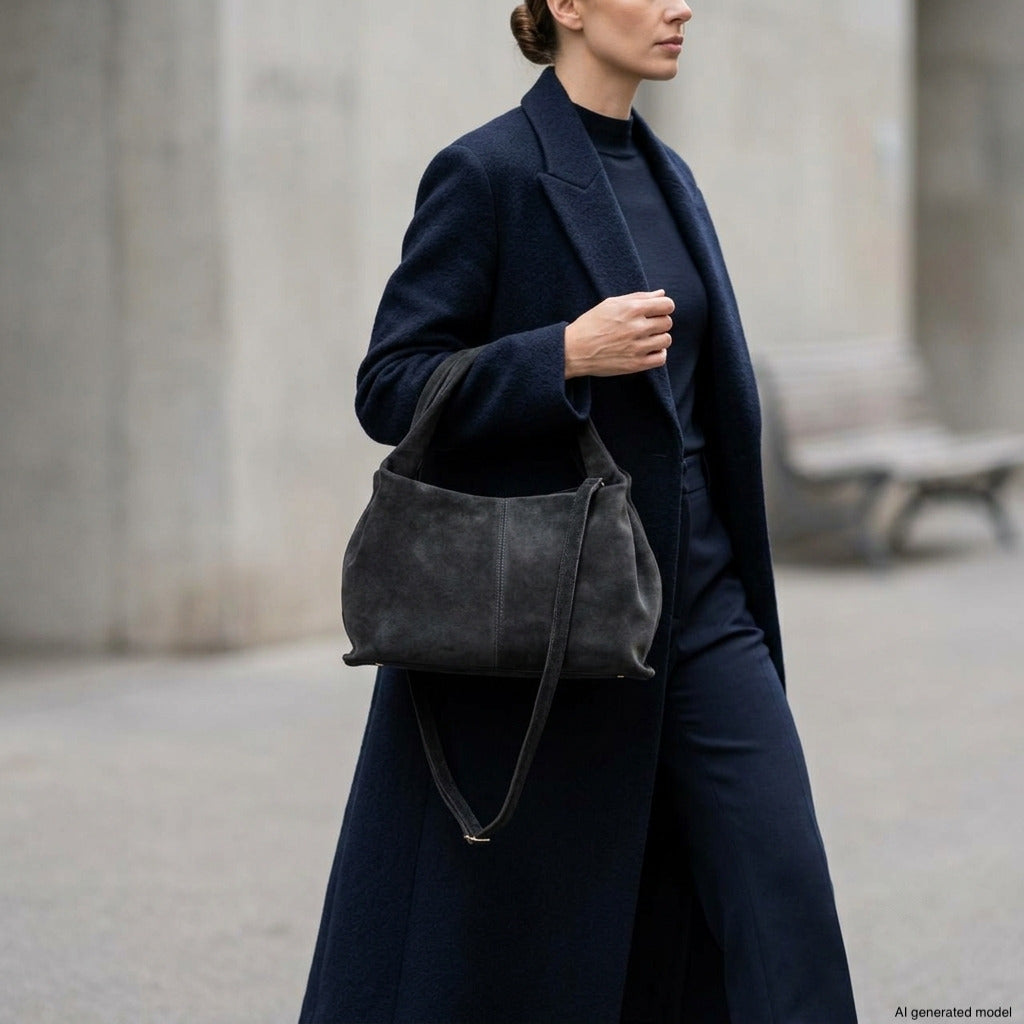 A person wearing a dark coat and pants carries a large handcrafted suede handbag, showcasing exquisite Italian craftsmanship while walking outdoors; a bench and concrete wall are visible in the background.