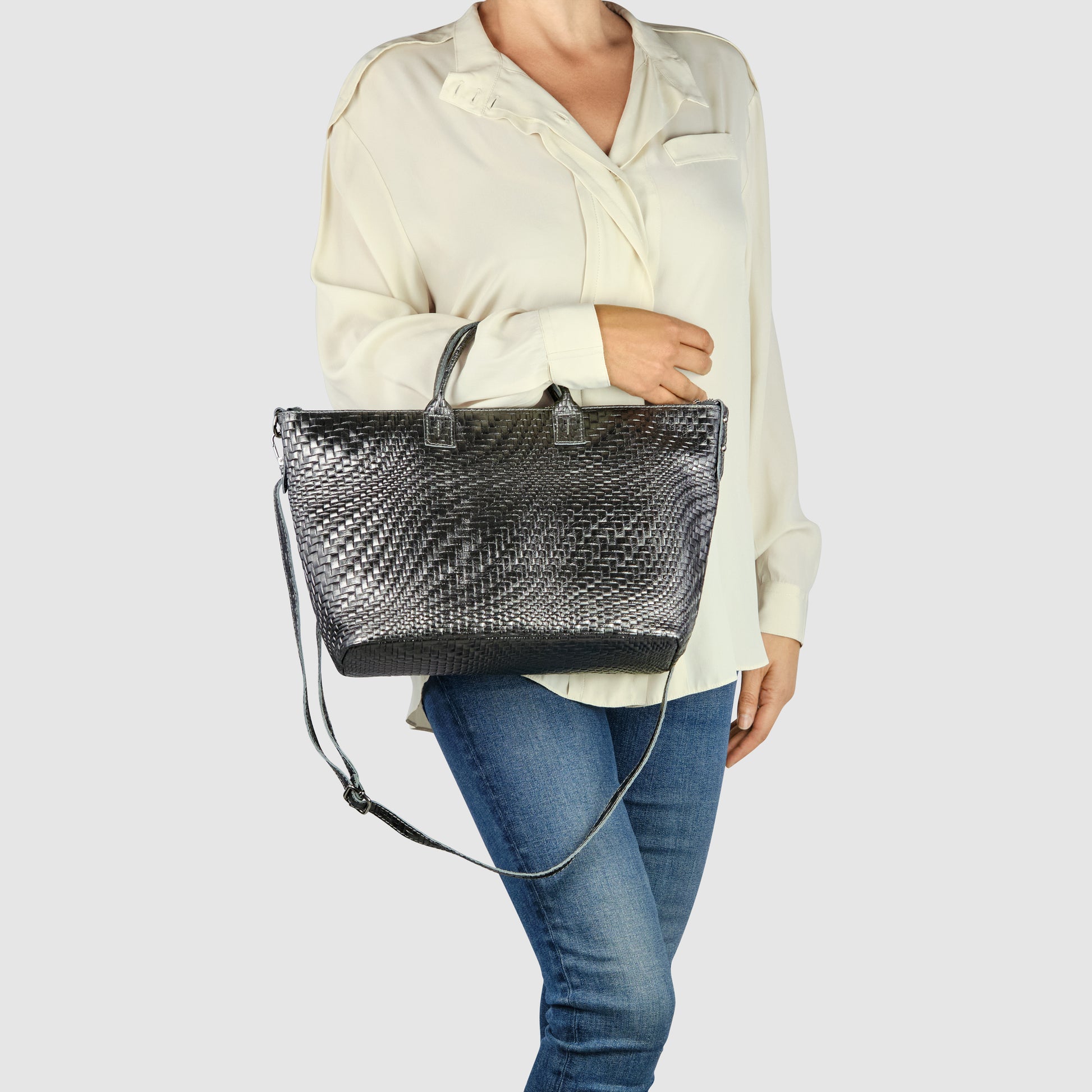 A woman wearing a cream blouse and blue jeans holds an Italian leather handbag with grid-embossed detailing and both handles and a long shoulder strap against a plain light background.