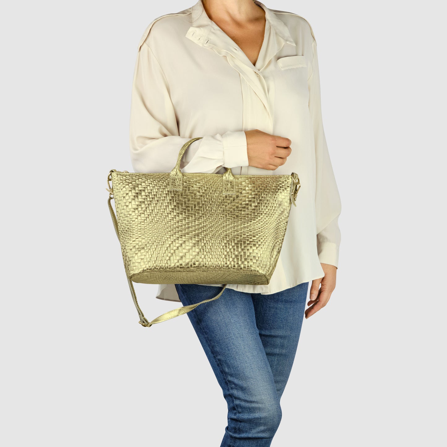 A person wearing a loose white blouse and blue jeans holds a chic grid-embossed purse with both a handle and a shoulder strap against a plain light background.