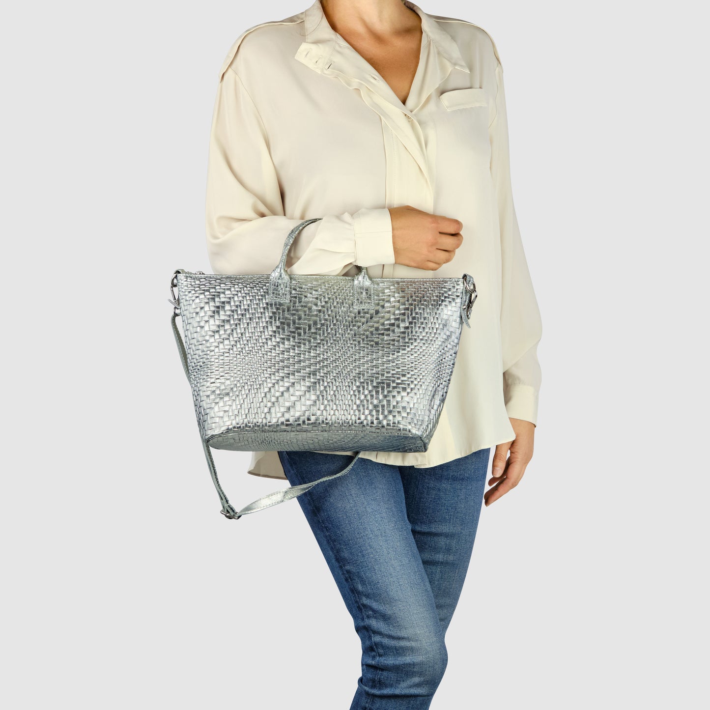A person wearing a loose cream blouse and blue jeans holds a stylish Italian leather handbag with both handles and a shoulder strap against a plain light background.