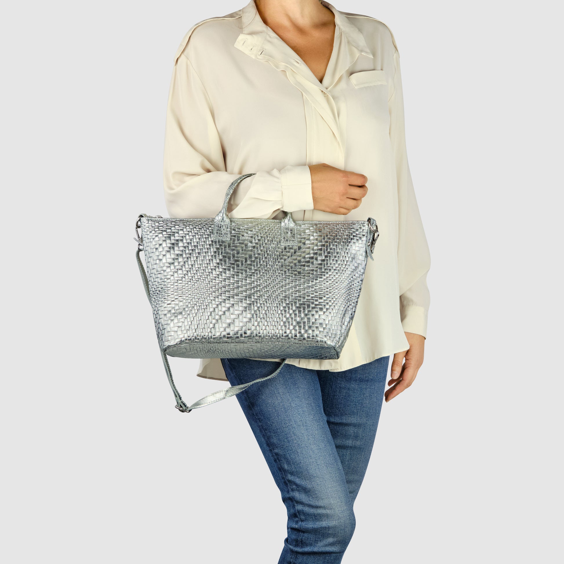 A person wearing a loose cream blouse and blue jeans holds a stylish Italian leather handbag with both handles and a shoulder strap against a plain light background.