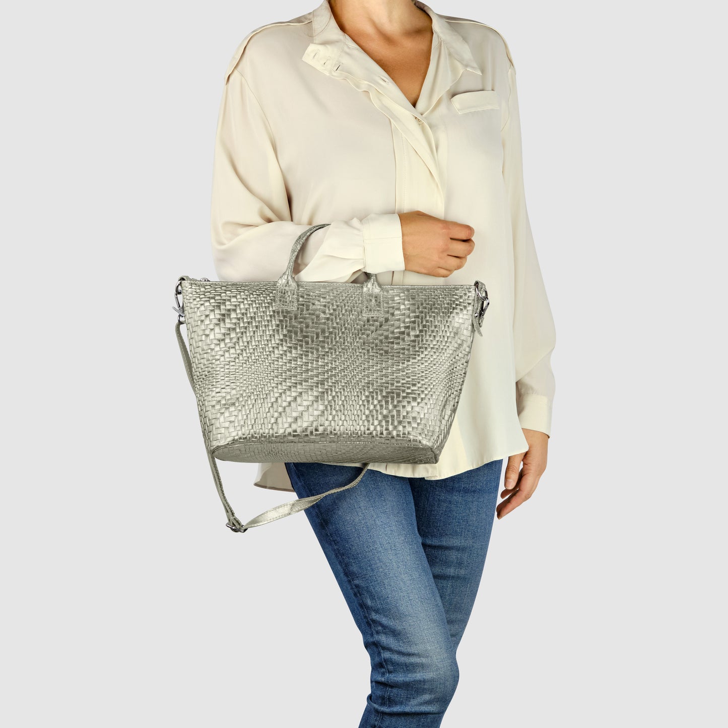 A woman wearing a cream blouse and blue jeans holds a large metallic finish purse with a shoulder strap, standing against a plain light grey background.