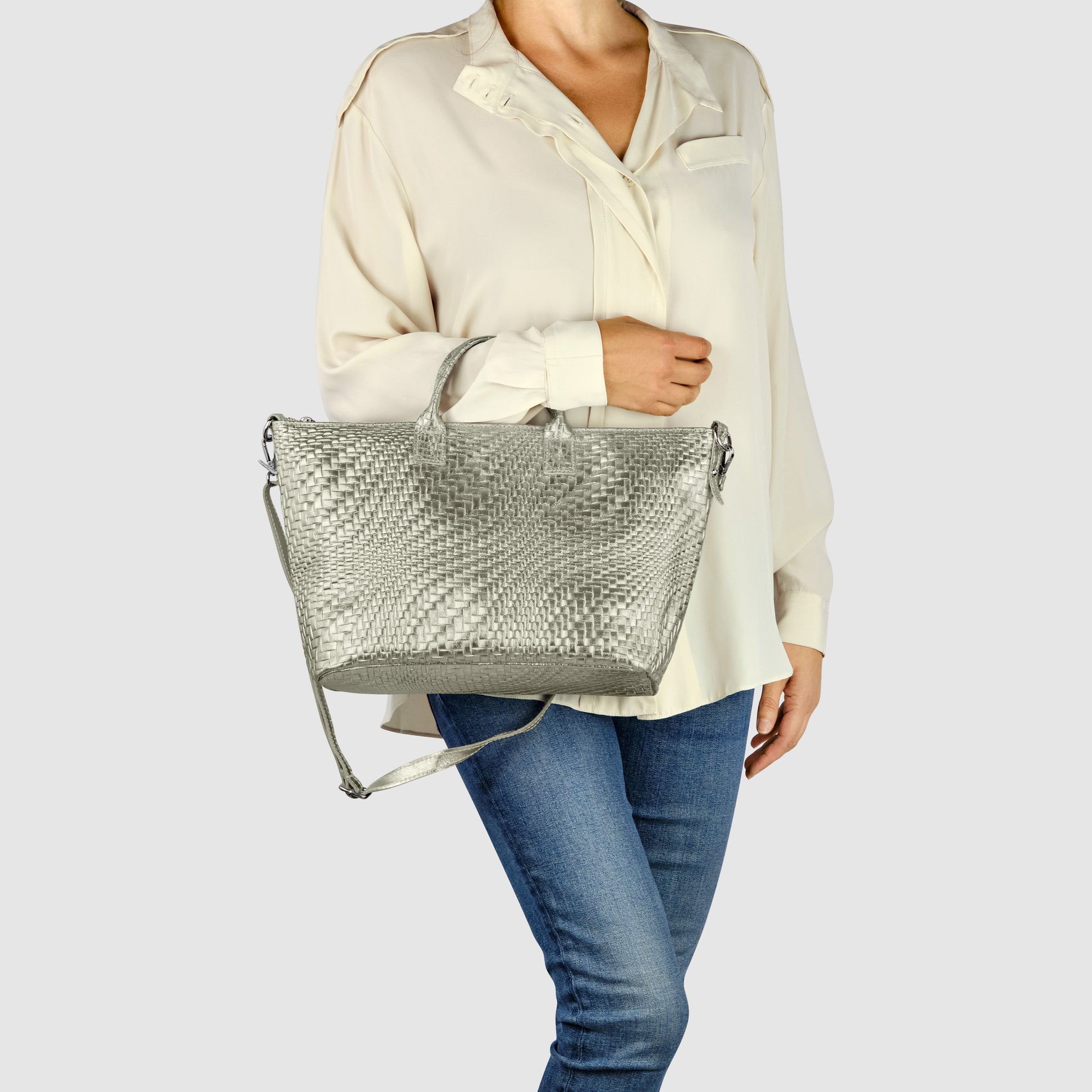 A woman wearing a cream blouse and blue jeans holds a large metallic finish purse with a shoulder strap, standing against a plain light grey background.