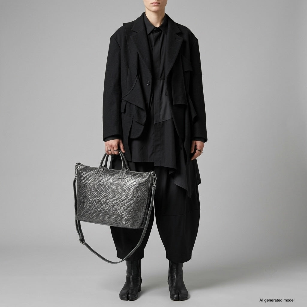A person wearing an oversized black suit with loose pants and a long coat stands holding a large Italian suede handbag. They are also wearing black ankle boots. The background is plain and light gray.