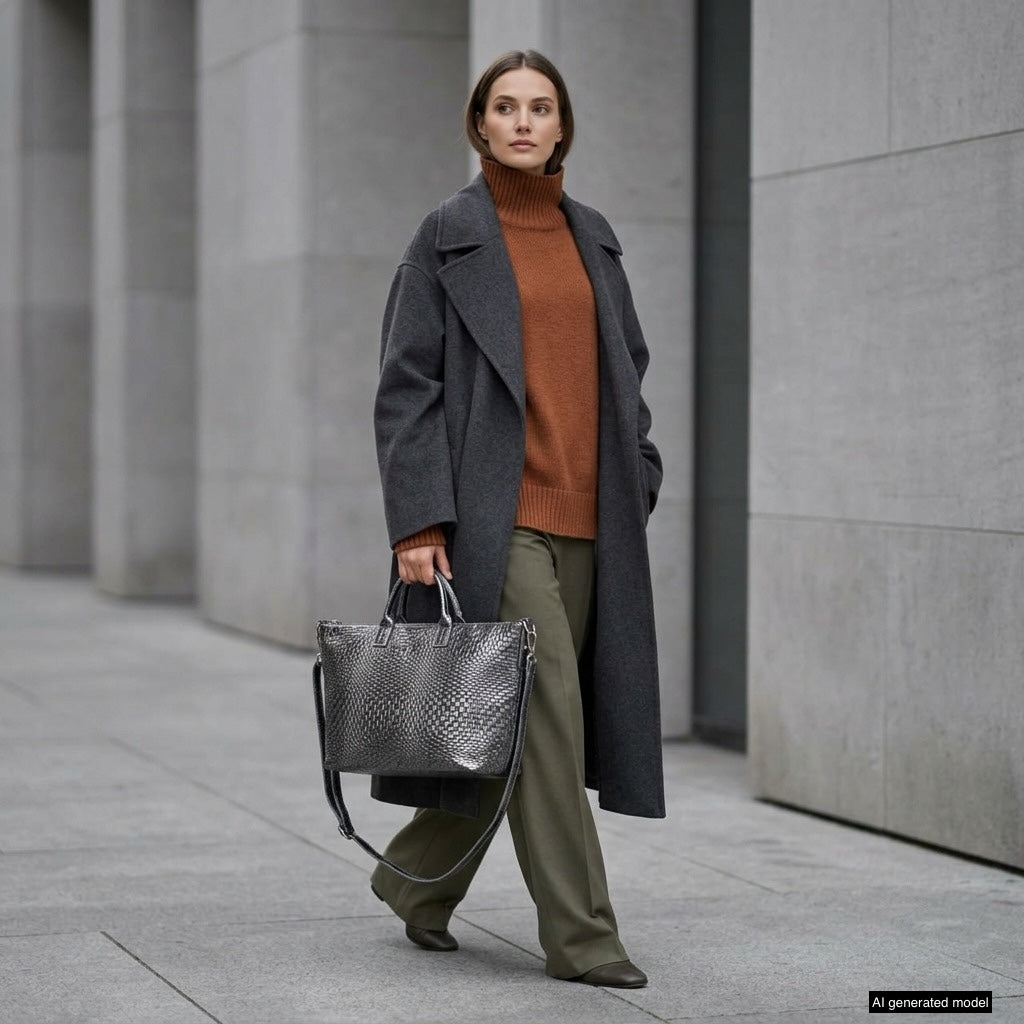 A woman in a gray overcoat, rust-colored turtleneck sweater, and olive-green pants walks on a city sidewalk, carrying an elegant Italian suede handbag. She stands against a backdrop of modern gray buildings.