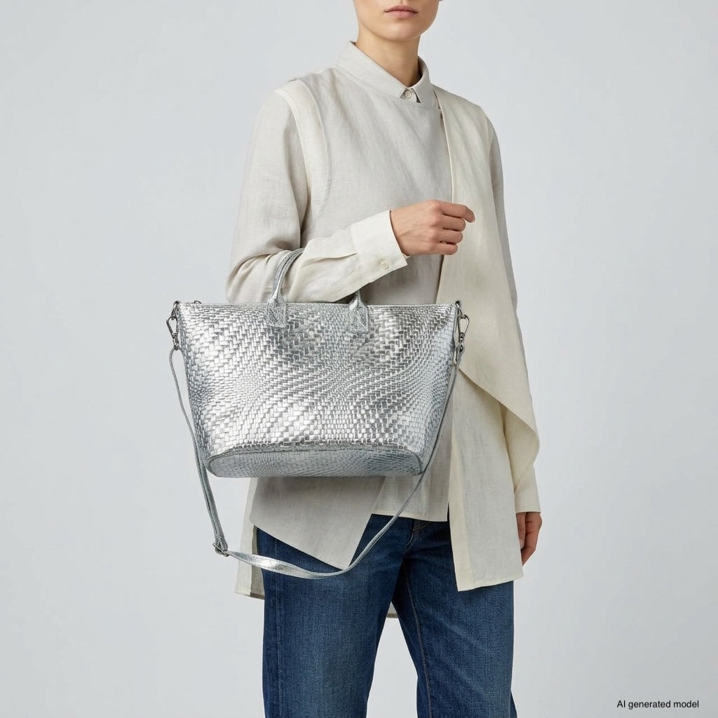 A person wearing a light beige blouse and blue jeans holds a metallic suede purse made in Italy, featuring a shiny silver textured finish with a shoulder strap, against a plain light gray background.
