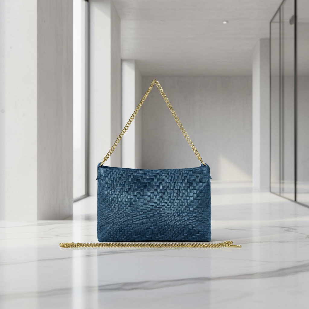 A blue woven suede leather handbag with a gold chain strap is displayed on a white marble surface in a modern, minimalistic room with large windows and concrete walls.