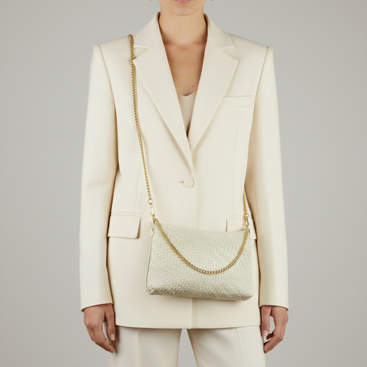 A person wearing a cream-colored suit stands facing forward, holding an Italian handcrafted bag with a gold chain strap over their shoulder. The textured, light-colored handbag stands out against the plain, light gray background.