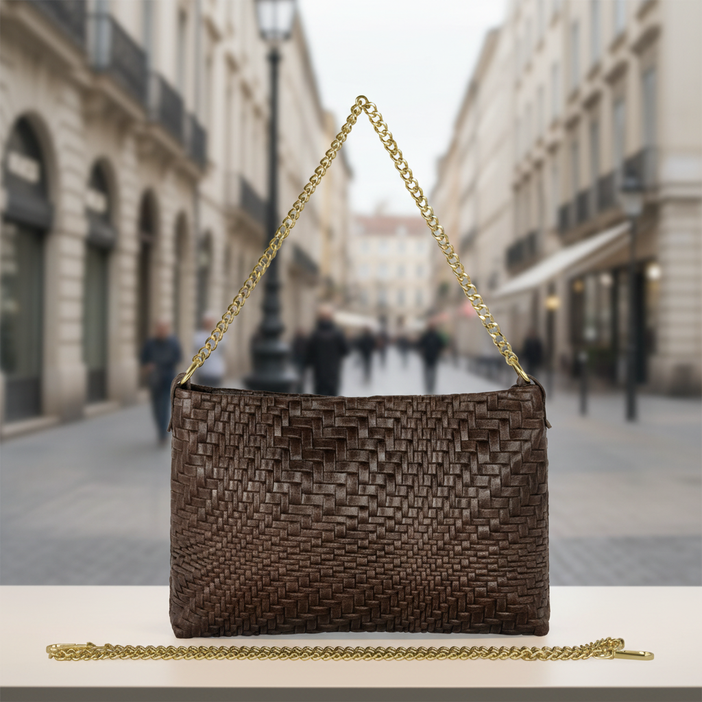 A dark brown Florentine leather handbag with a gold chain strap is displayed on a white surface. A matching chain lies in front. The background shows a blurred city street with classic buildings and a few people walking.