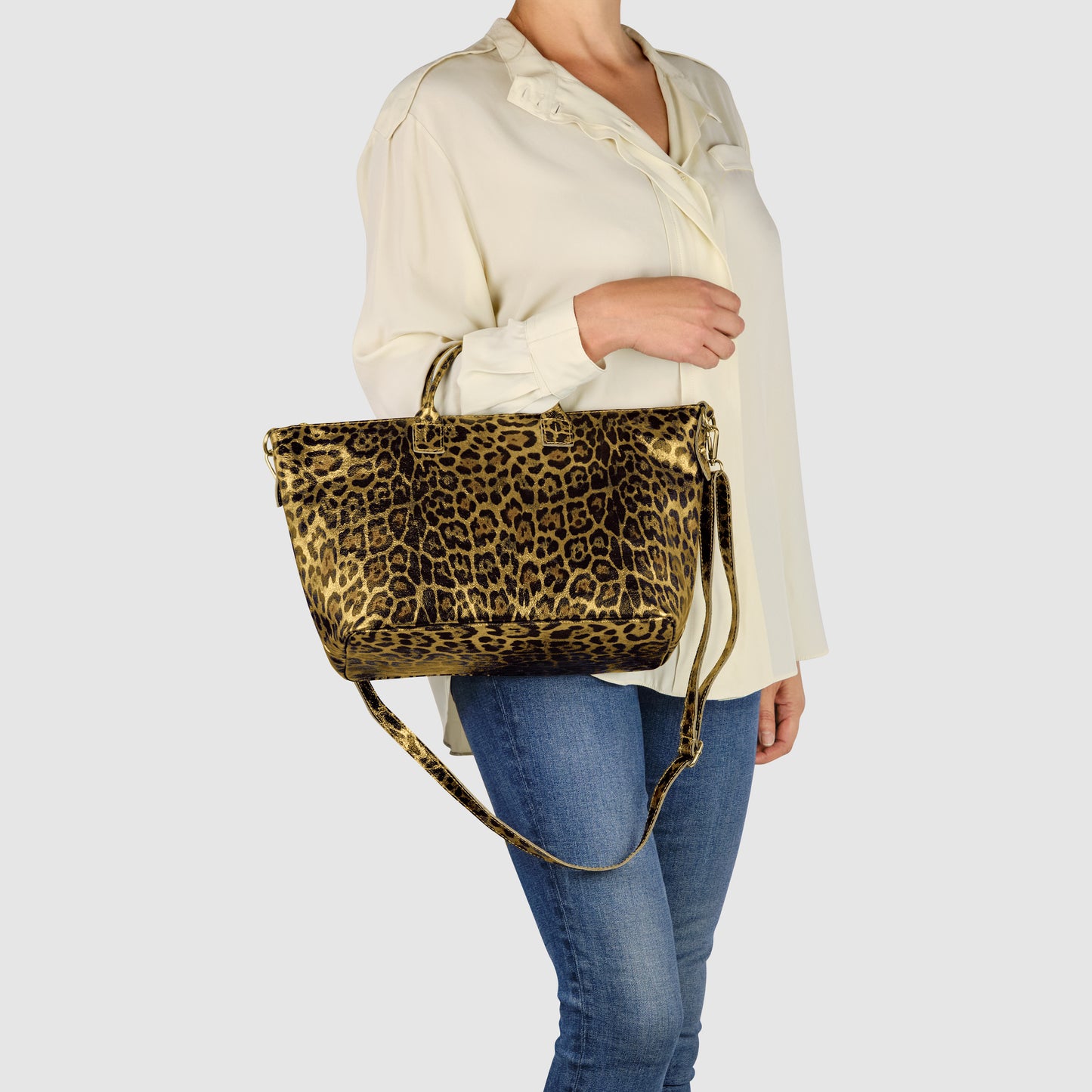 A person wearing a cream blouse and blue jeans holds a metallic leopard print handbag with both short handles and a long shoulder strap against a plain light background.