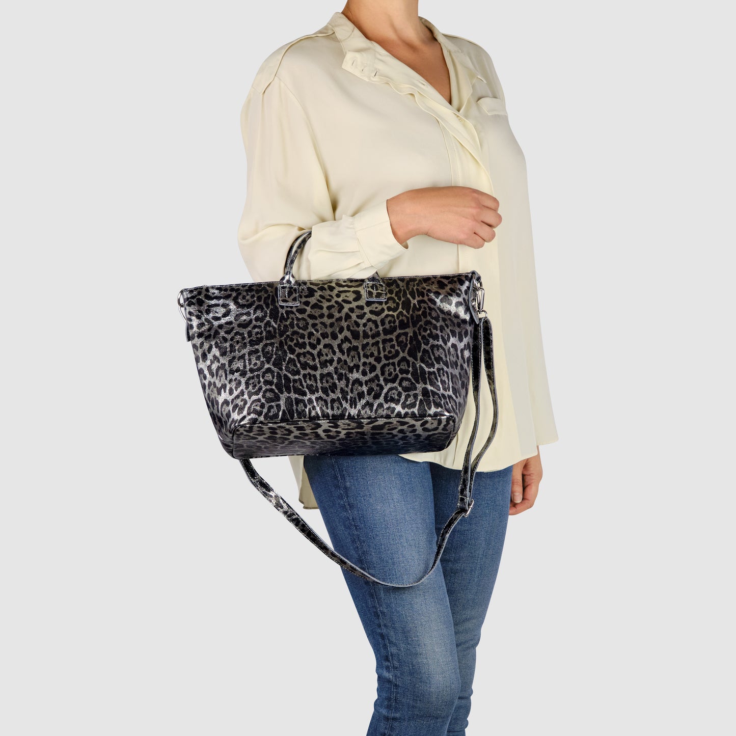 A person wearing a cream blouse and blue jeans holds a TAKEASY metallic leopard print handbag with both short handles and a long shoulder strap against a plain light background.