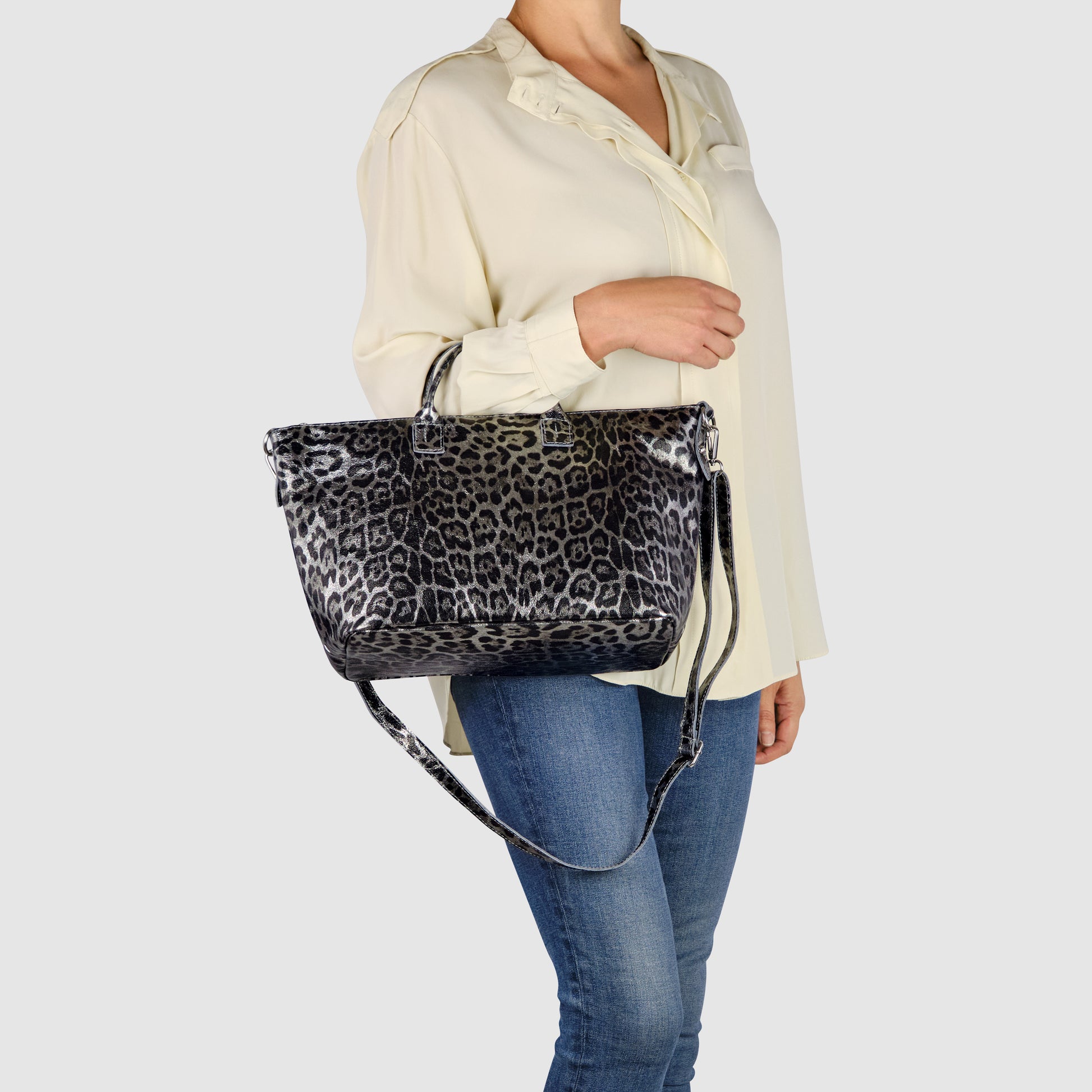 A person wearing a cream blouse and blue jeans holds a TAKEASY metallic leopard print handbag with both short handles and a long shoulder strap against a plain light background.