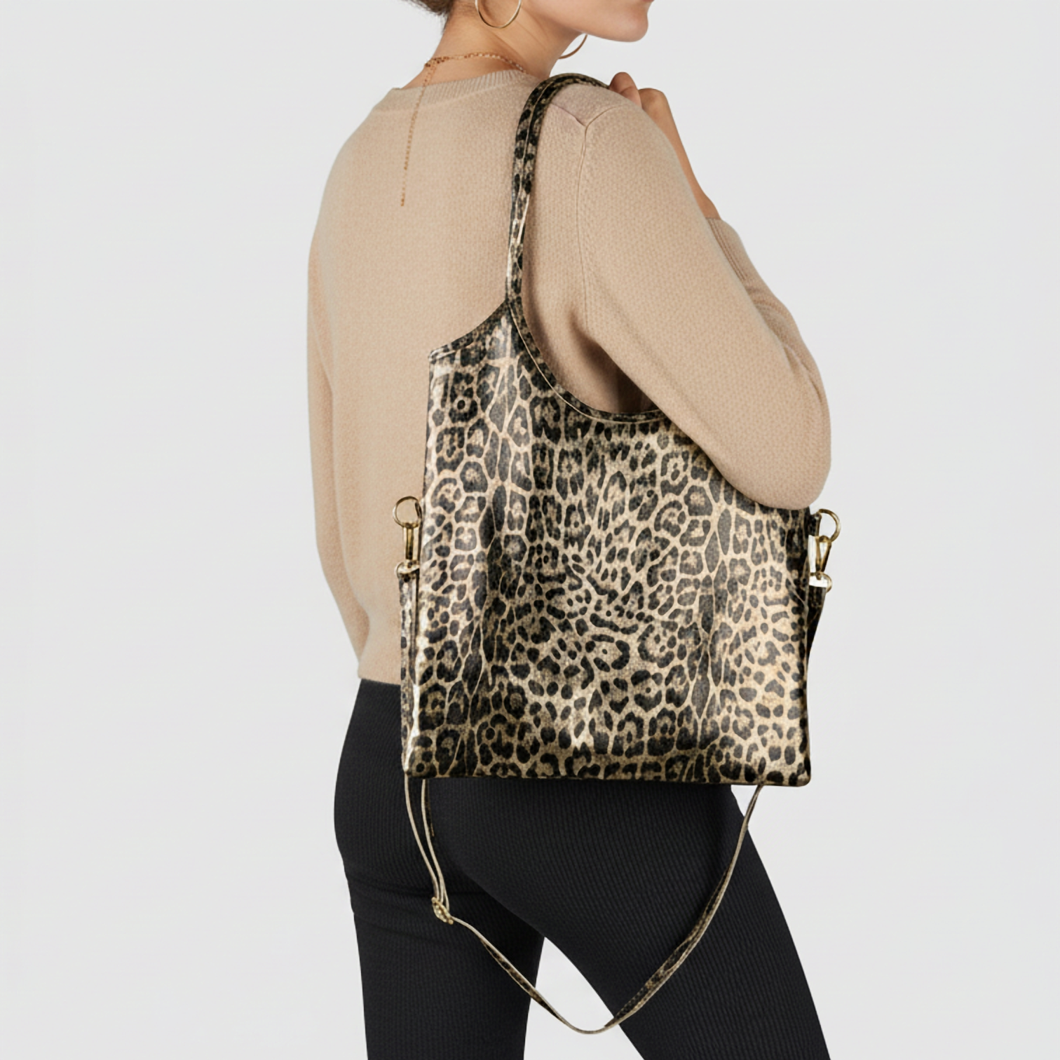 A woman in a beige long-sleeve top and black pants carries an Italian leather bag with leopard embossing and gold hardware over her shoulder, standing against a plain light background.