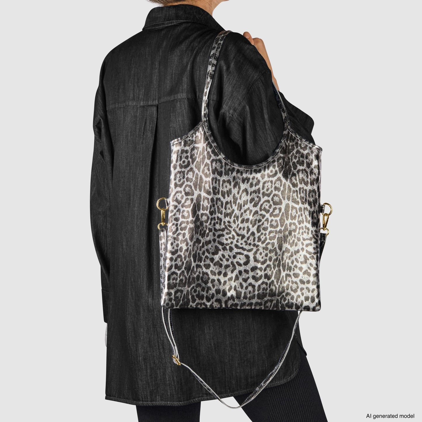 A person in a black jacket holds a large, handmade suede bag with gray-and-white leopard print, gold hardware, and a detachable strap against a plain white background.