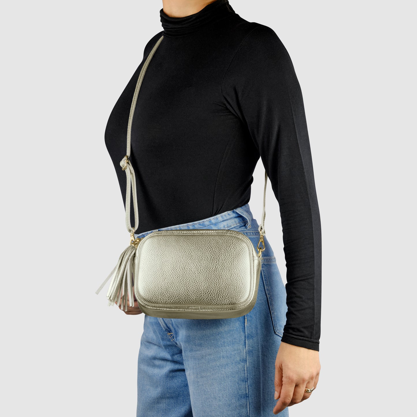 A person wearing a black long-sleeve turtleneck and blue jeans carries a luster calfskin crossbody handbag with a tassel detail against a plain light background.