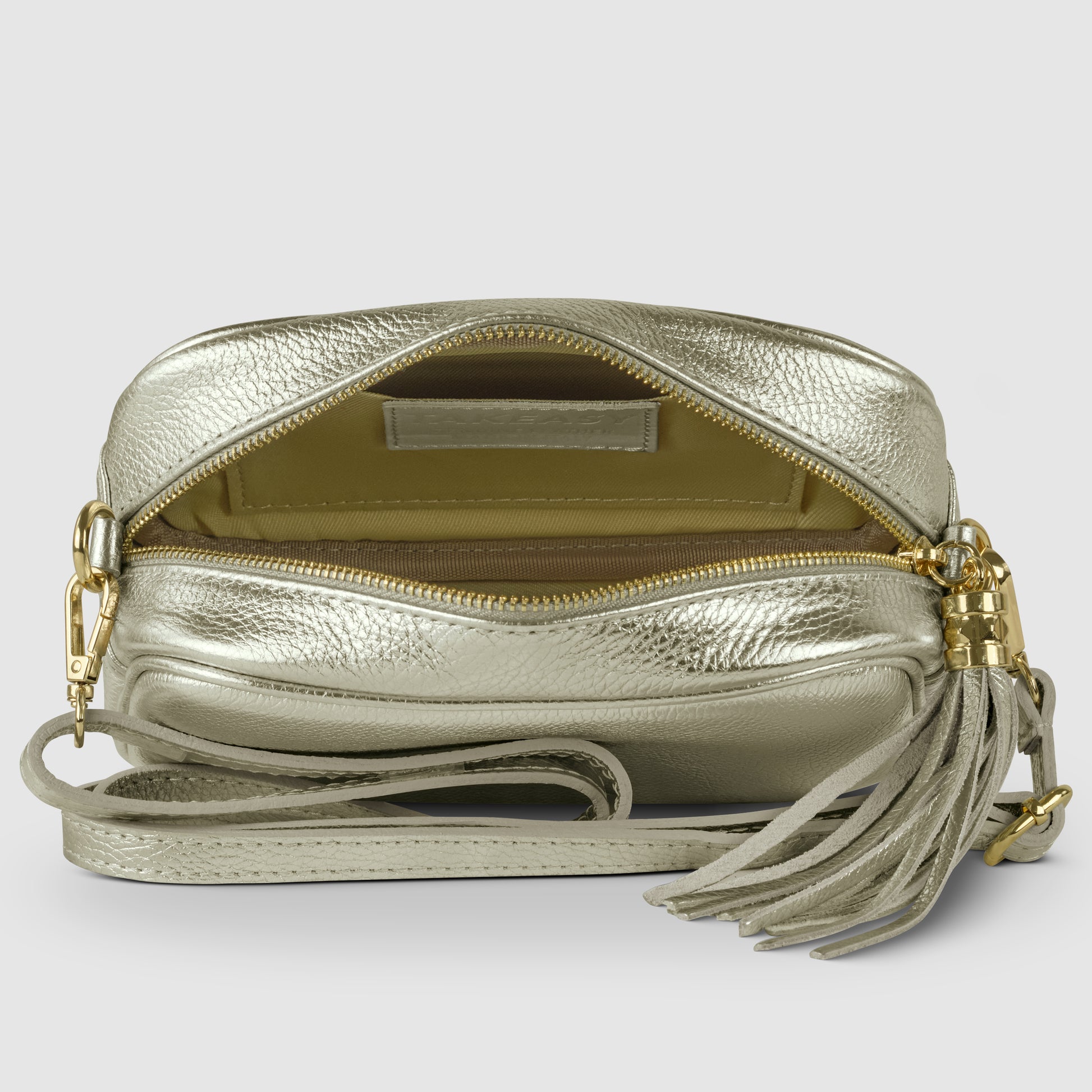 A metallic gold luster calfskin crossbody handbag with a tassel zipper pull is shown open, revealing a beige interior and an inner pocket. The bag features a matching strap and gold hardware.