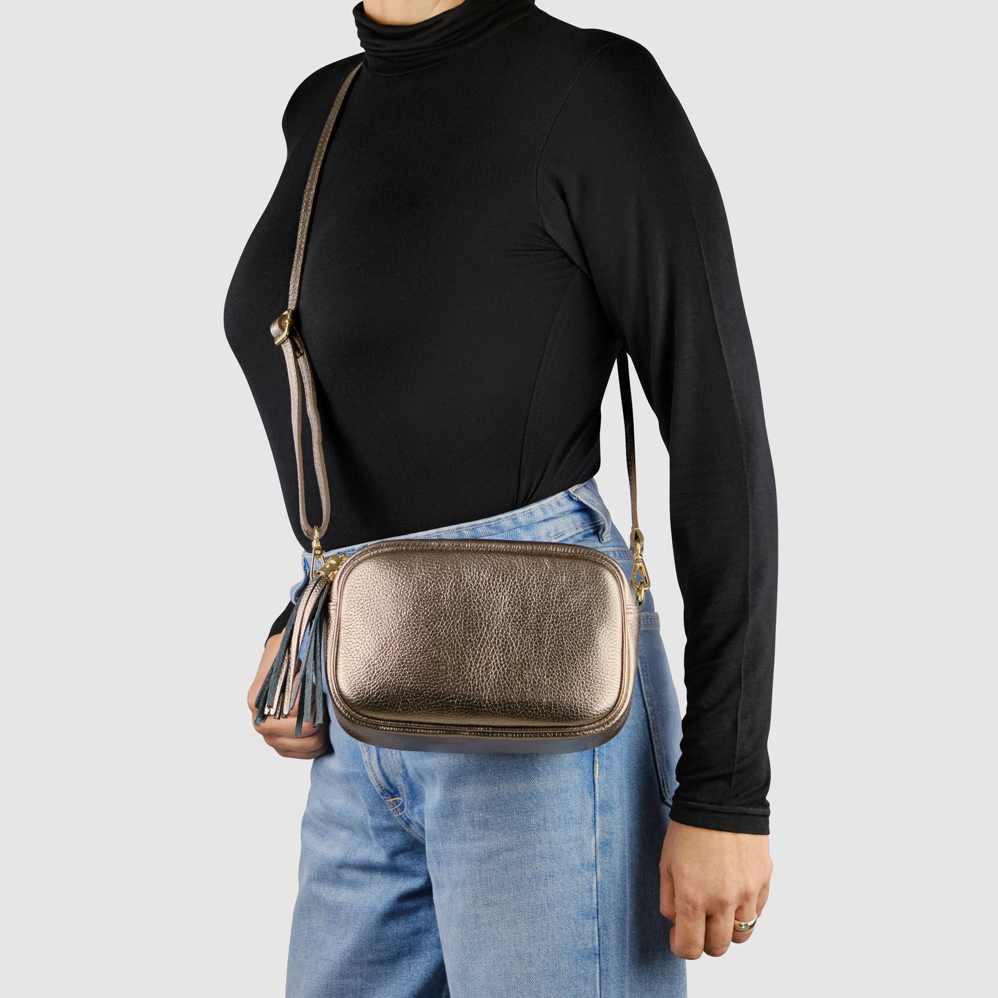 A person wearing a black turtleneck and blue jeans models a metallic luster calfskin crossbody handbag with a tassel detail, worn over the shoulder against a plain light background.