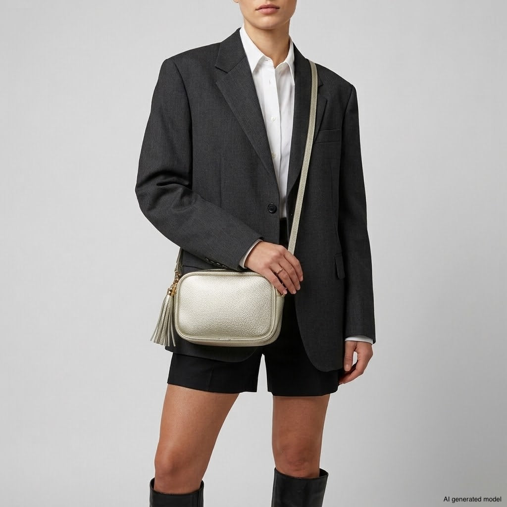 A person wearing a dark blazer, white shirt, black shorts, and knee-high boots holds a metallic finish purse with a tassel. The background is plain and light-colored.