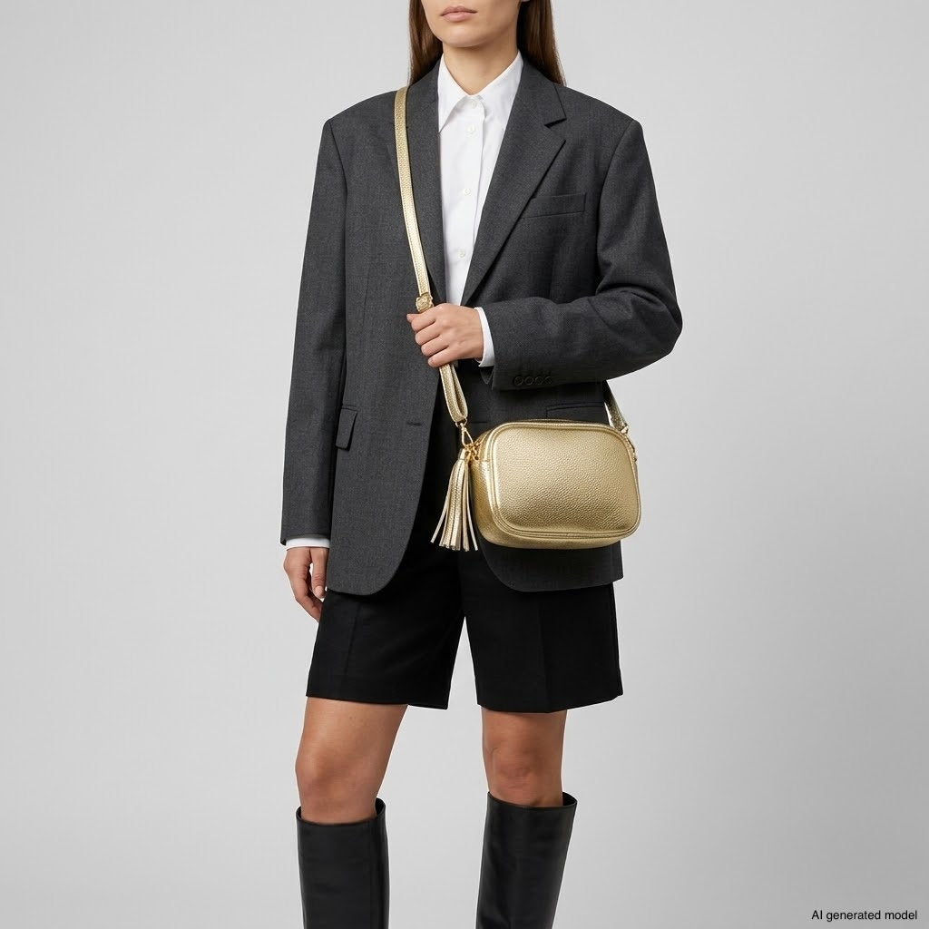 A person in a dark blazer, white shirt, and black shorts stands with a metallic finish purse and tall black boots against a plain background.