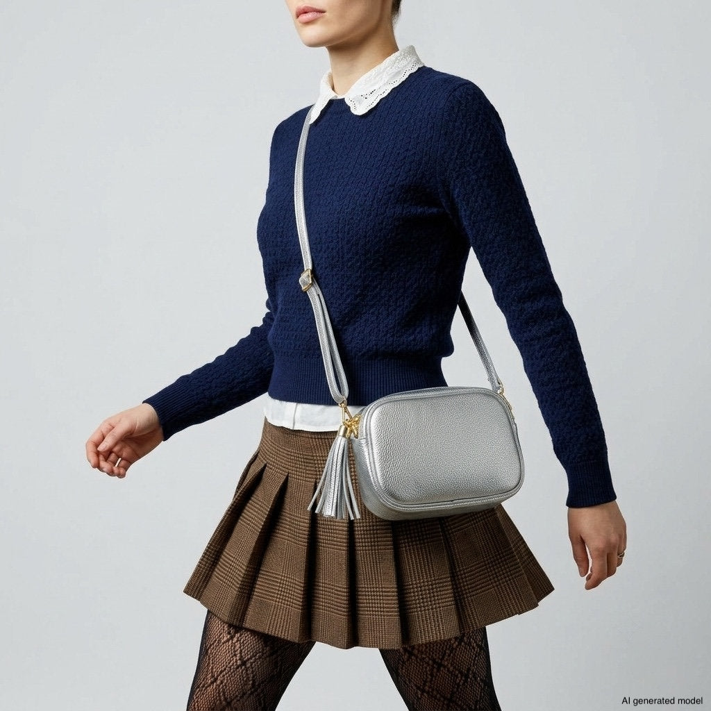A person wearing a navy sweater, white collared shirt, brown pleated skirt, patterned tights, and carrying a metallic finish purse walks against a plain background.