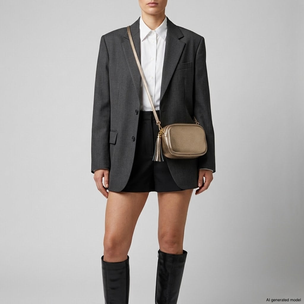 A person wearing a gray blazer, white shirt, black shorts, knee-high black boots, and an Italian full-grain leather gold crossbody bag stands against a plain background.