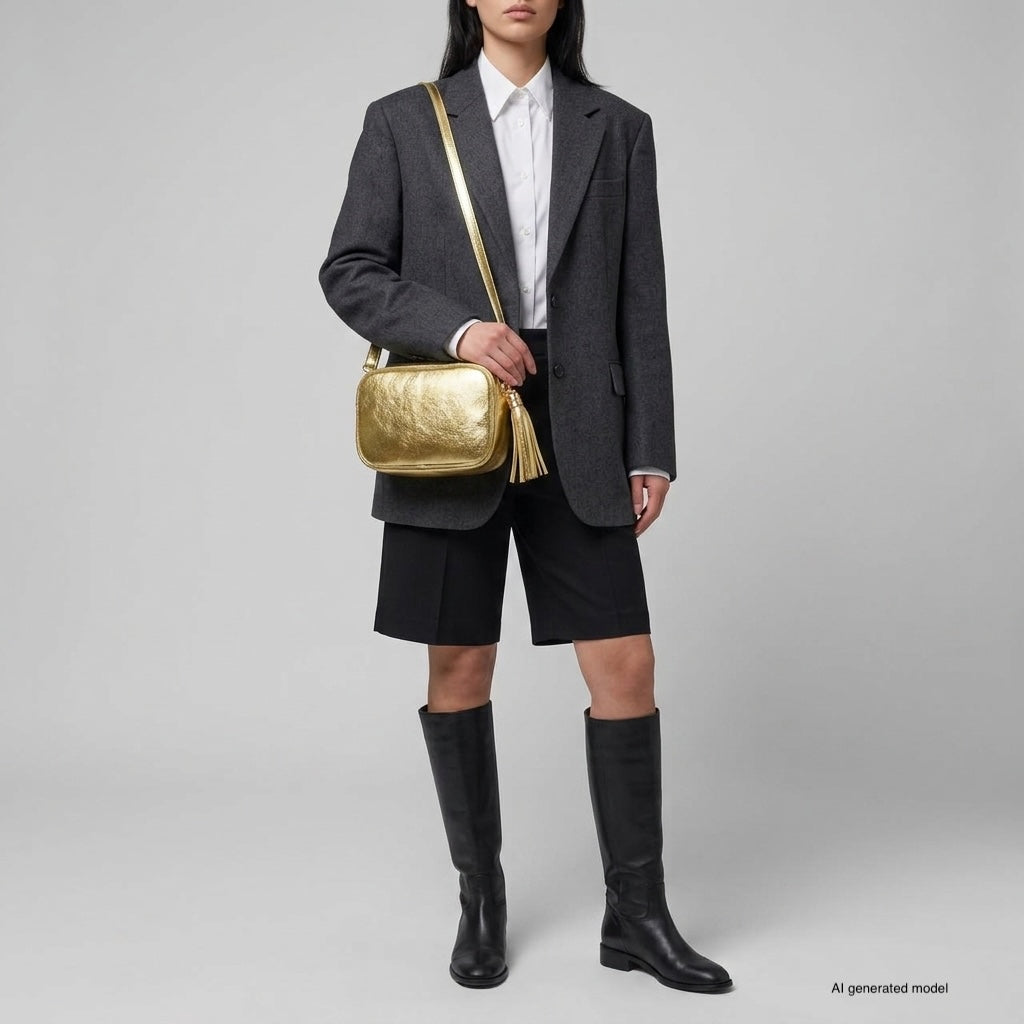 A person wearing a dark gray blazer, white shirt, black knee-length shorts, and knee-high black boots stands against a plain background, holding a compact rectangular handbag crafted from shiny Italian leather.