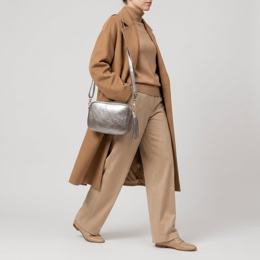 A person wearing a brown long coat, tan sweater, wide-leg beige trousers, and tan shoes walks while carrying a compact rectangular handbag showcasing Italian craftsmanship against a plain light background.