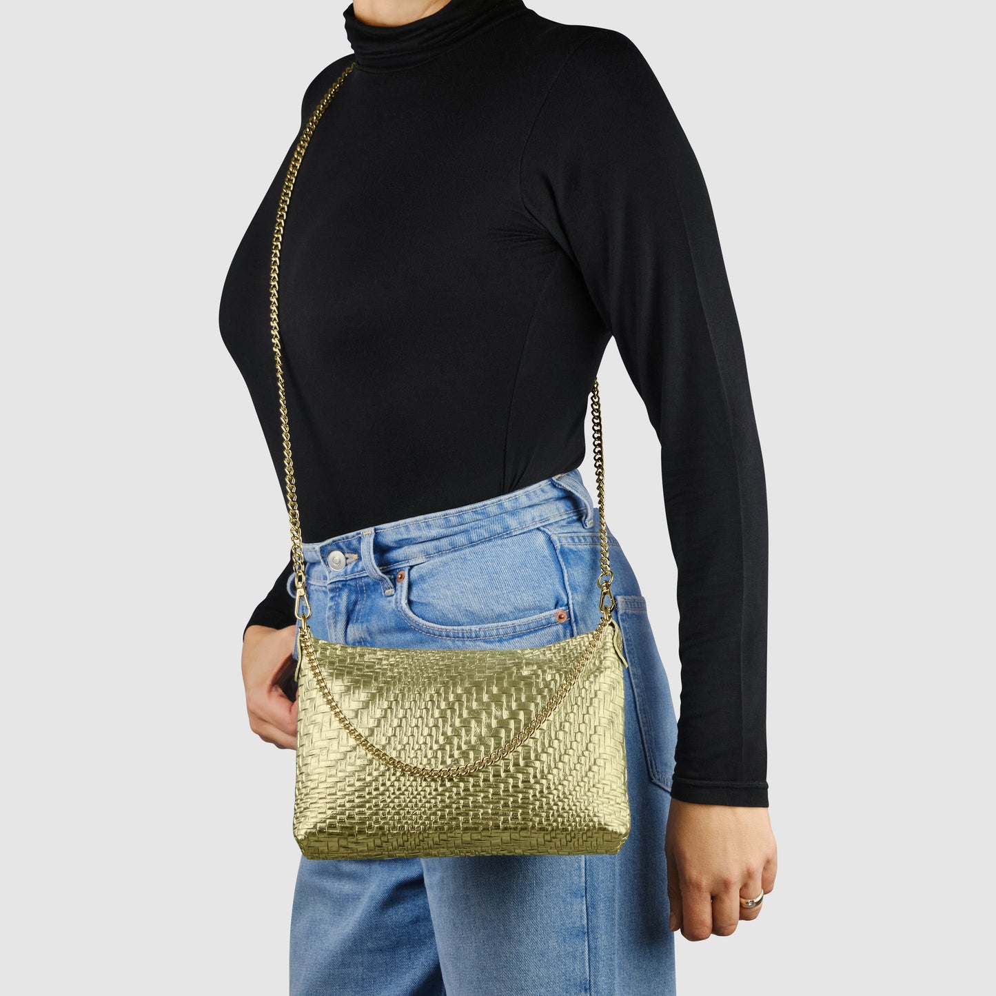 A person in a black long-sleeve turtleneck and blue jeans holds an Italian handcrafted handbag—a gold metallic-woven leather bag with a chain strap—posed against a plain light gray background.
