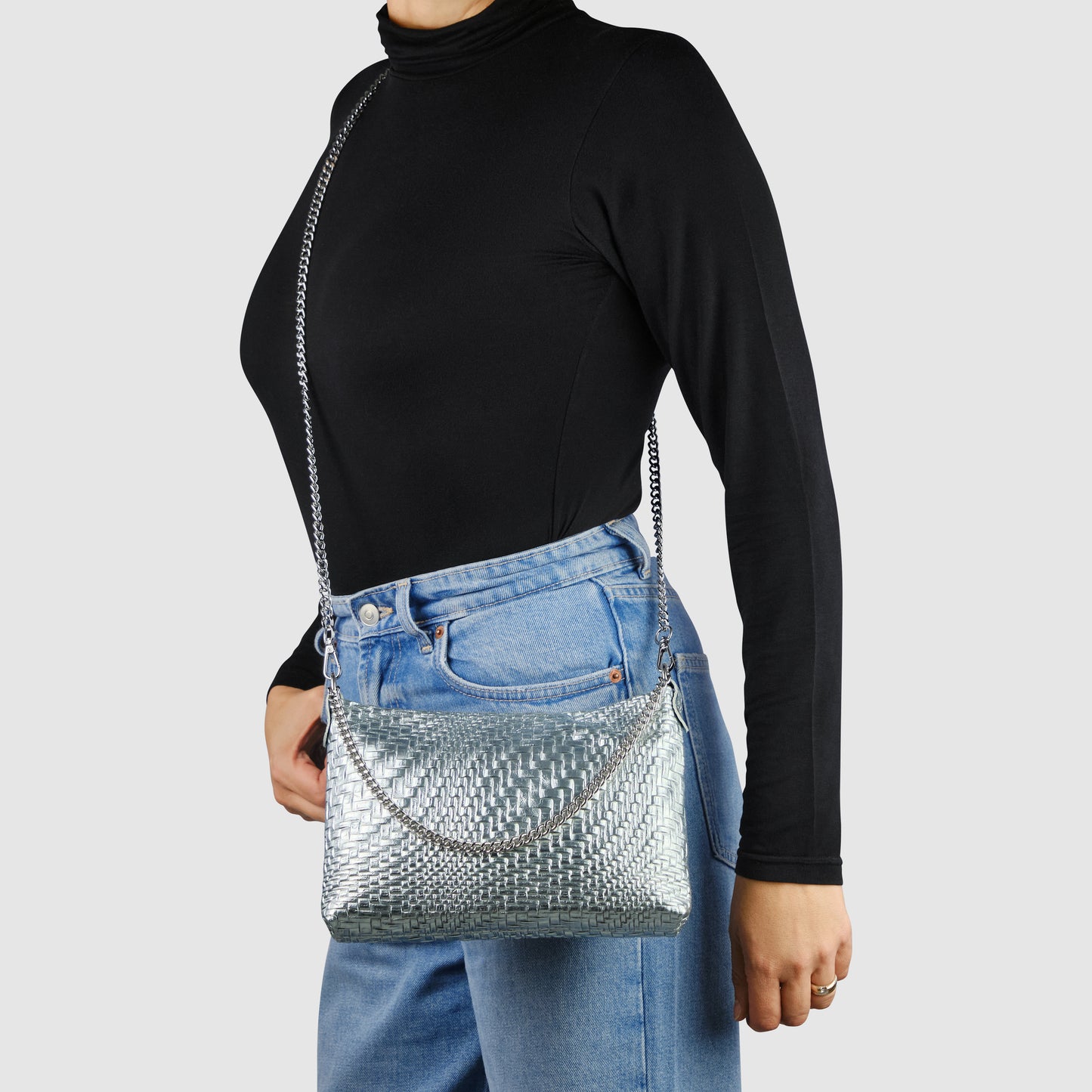 A person wearing a black turtleneck and blue jeans carries a metallic-woven leather bag with a chain strap, showcasing Florentine leathercraft against a plain light gray background.