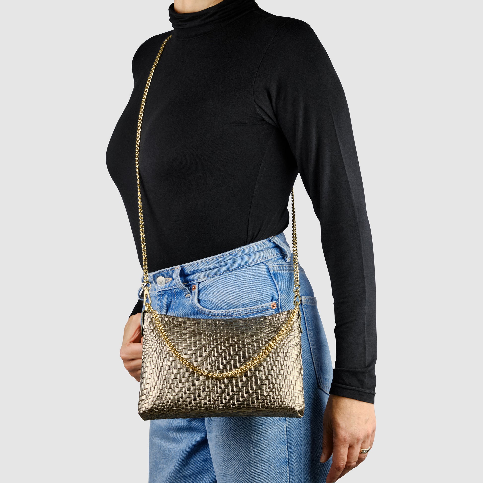 A person in a black turtleneck and blue jeans carries a metallic-woven leather bag with a chain strap, standing against a plain light gray background.