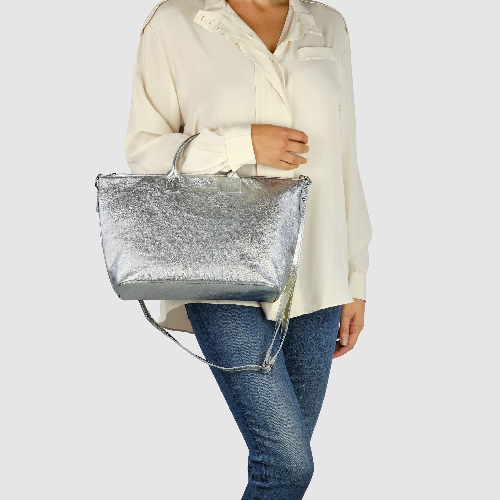 A woman wearing a cream-colored blouse and blue jeans holds a sleek Italian handbag with both handles and a shoulder strap against a plain light background.