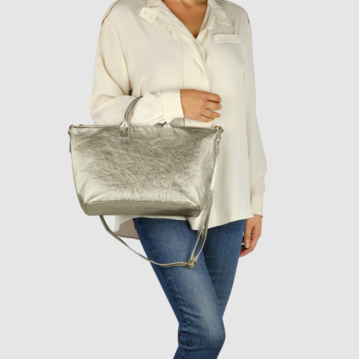 A woman wearing a loose white blouse and blue jeans holds a striking Italian handbag with a metallic finish and shoulder strap against a plain gray background.