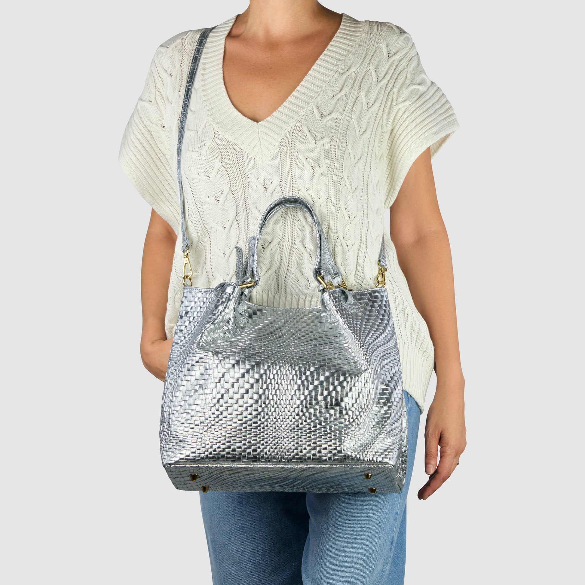 A woman in a cream short-sleeved knitted sweater and blue jeans carries a large women's handbag with metallic silver texture, featuring both top handles and a shoulder strap. The background is plain light gray.