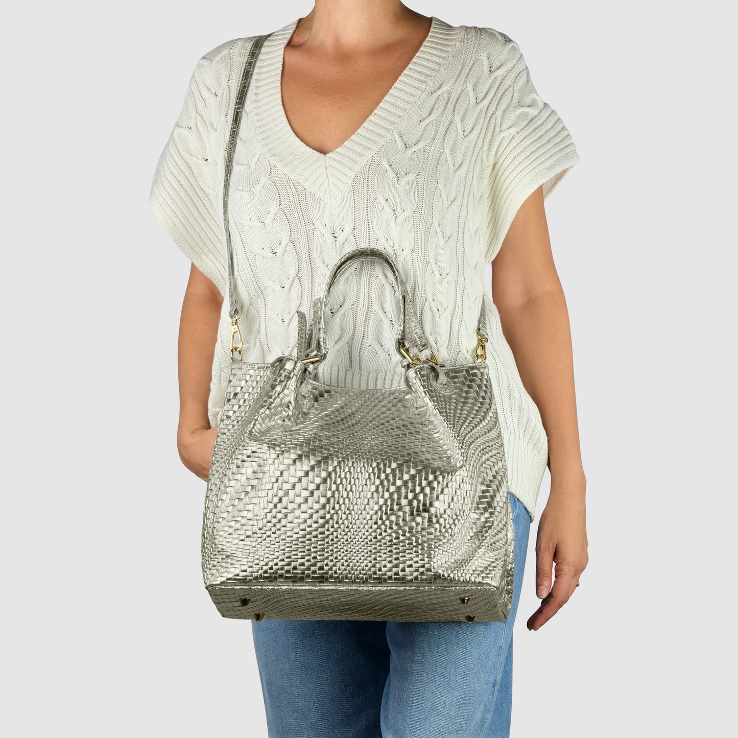 A person wearing a cream cable-knit short-sleeve sweater and blue jeans carries a large metallic silver handcrafted designer bag with top handles and a shoulder strap.