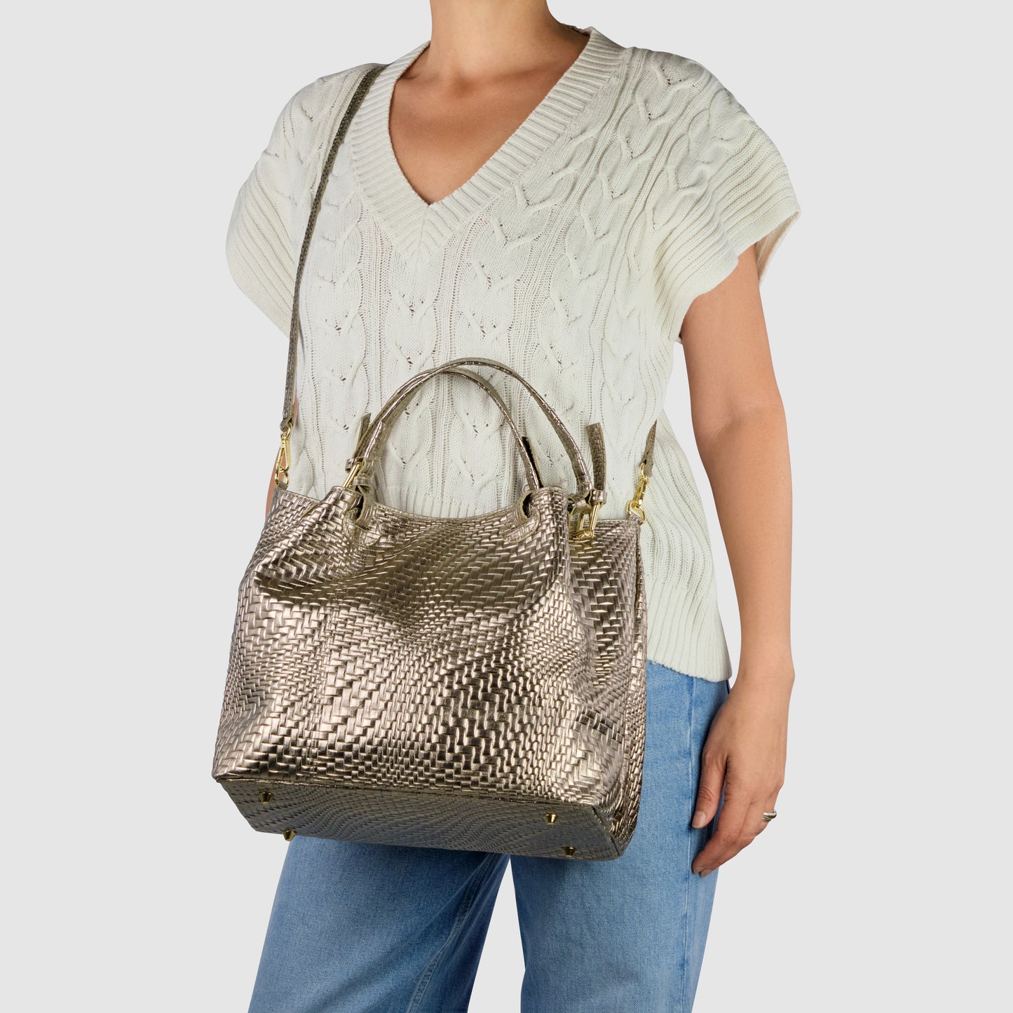A woman in a white knit sweater and blue jeans carries a women's designer bag with both handles and a shoulder strap. The metallic textured handbag features a woven pattern, gold hardware, and showcases modern elegance.