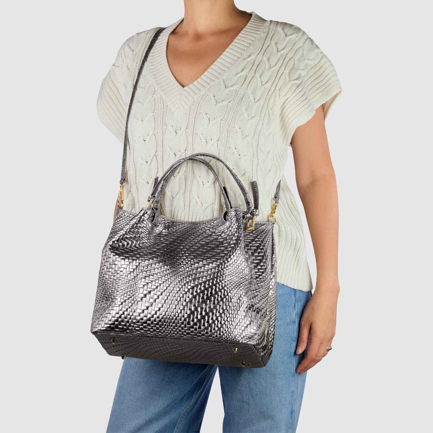 A person wearing a short-sleeved, cream cable-knit sweater and blue jeans carries an Italian leather handbag with a metallic silver texture, featuring both a shoulder strap and two handles. The background is plain light gray.