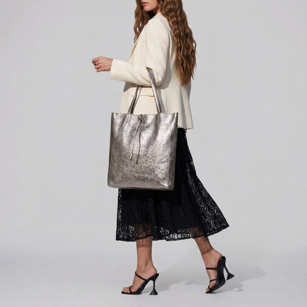 A woman with long wavy hair wears a cream blazer, black lace skirt, and black heeled sandals, carrying a handmade bag inspired by real Tuscan craft while walking against a plain light background.