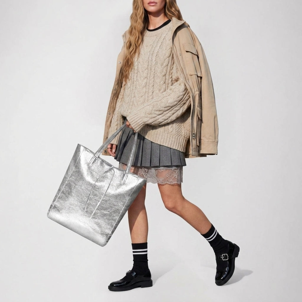 A woman wearing a beige cable-knit sweater over a tan jacket, gray pleated skirt, black loafers, and black socks with white stripes carries a handmade bag, perfectly complementing her long wavy hair and chic ensemble.