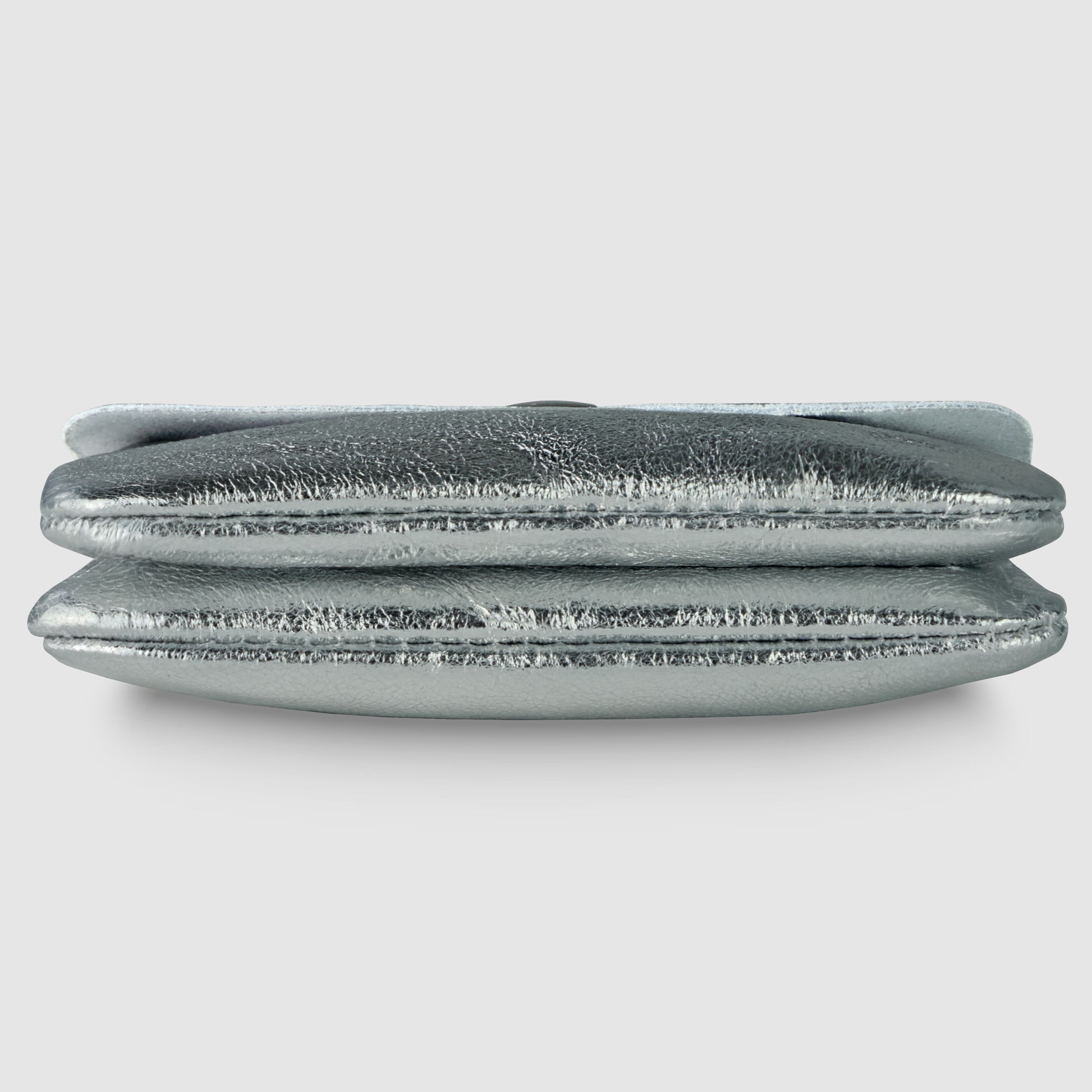 A metallic silver Gleam Suede Leather clutch purse is shown lying flat, viewed from the side, with a soft, textured surface and a fold-over design that reflects quiet luxury against a plain light background.