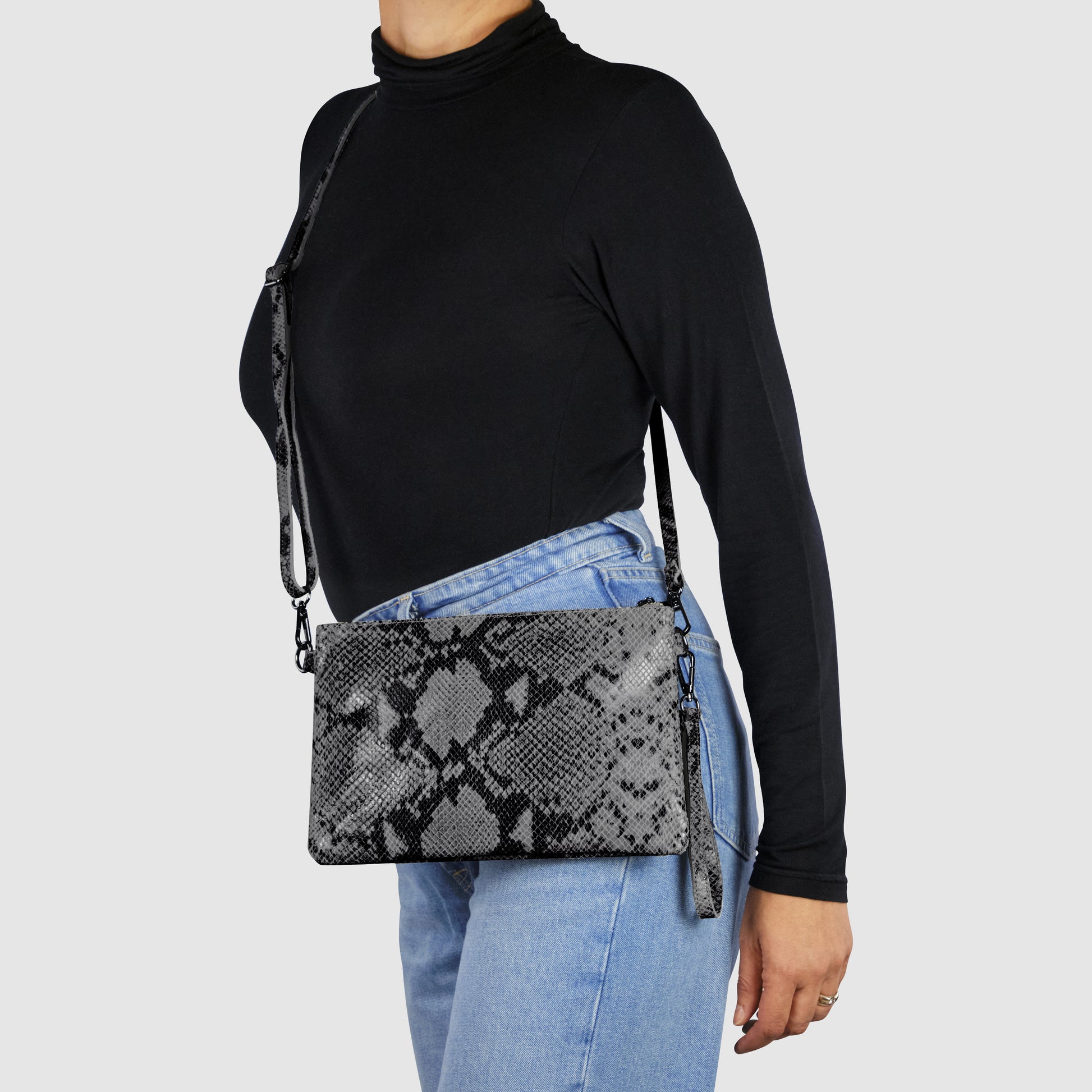 A person in a black turtleneck and blue jeans is wearing an Italian handcrafted python suede bag with a matching strap, standing against a plain light gray background.