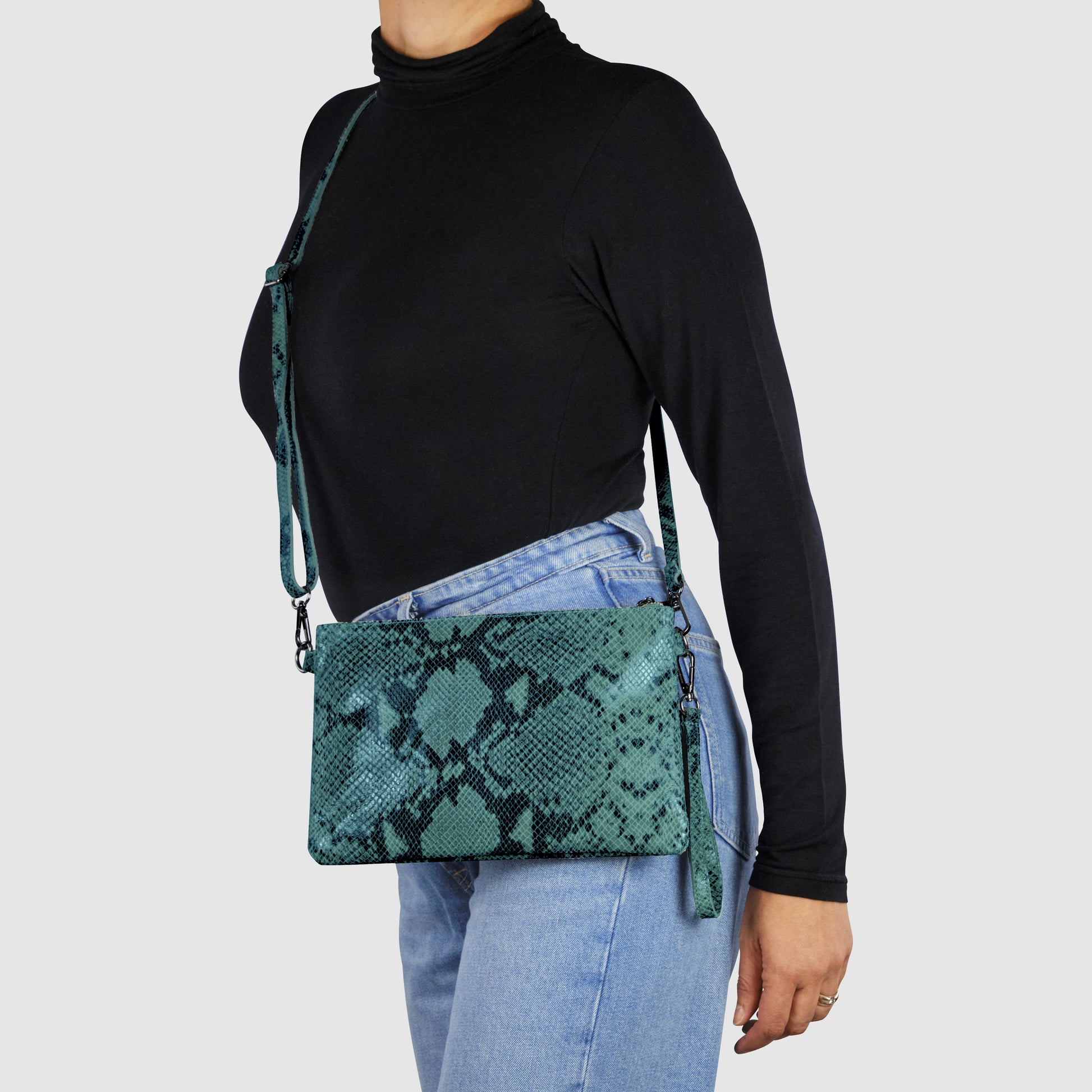 A person wearing a black turtleneck and light blue jeans is modeling an Italian handcrafted python suede bag with a matching strap, standing against a plain light gray background.