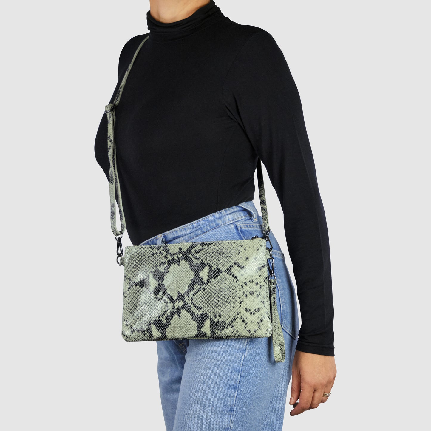 A person in a black turtleneck and light blue jeans carries an Italian handcrafted python suede bag with a green snakeskin pattern and matching strap against a plain light background.