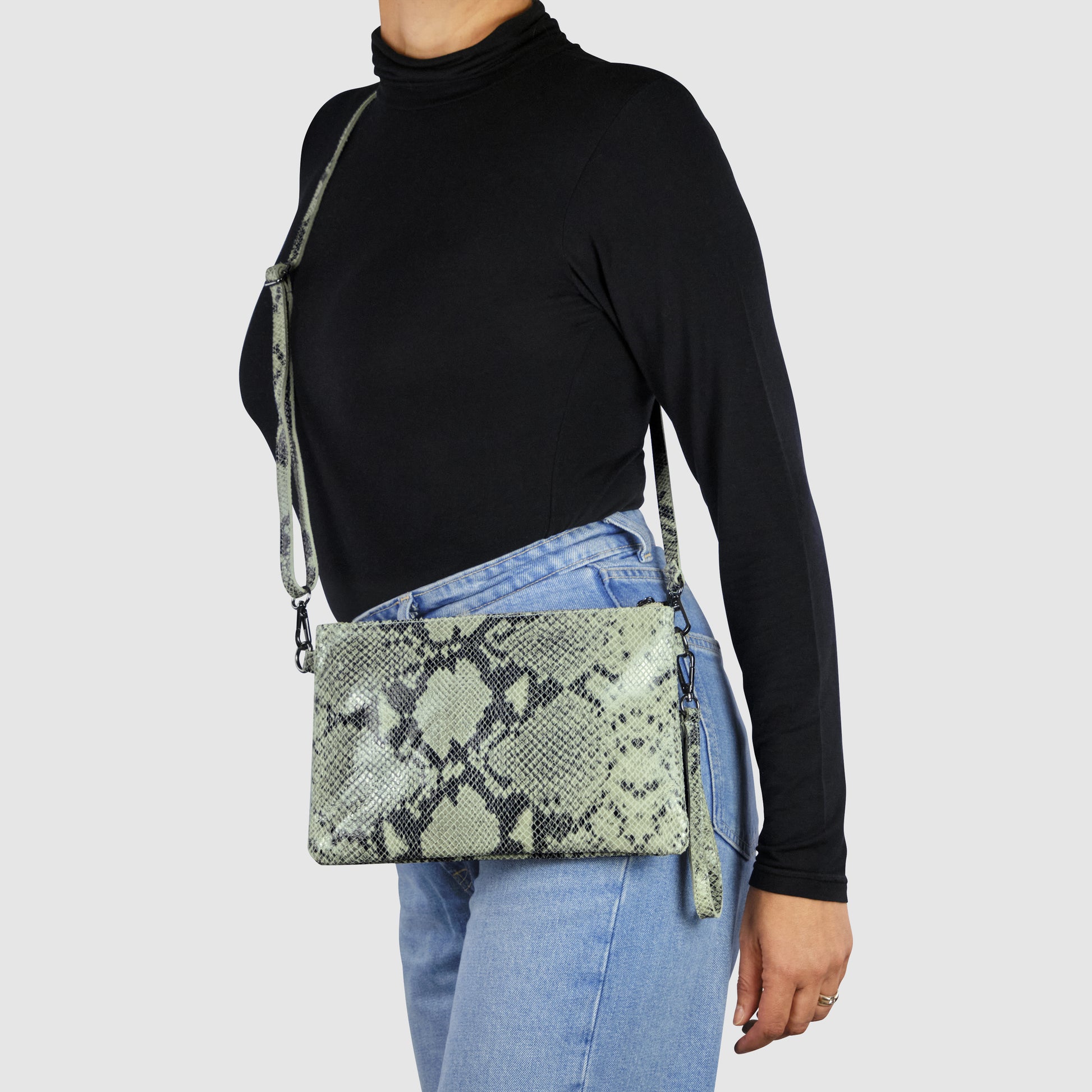 A person in a black turtleneck and light blue jeans carries an Italian handcrafted python suede bag with a green snakeskin pattern and matching strap against a plain light background.