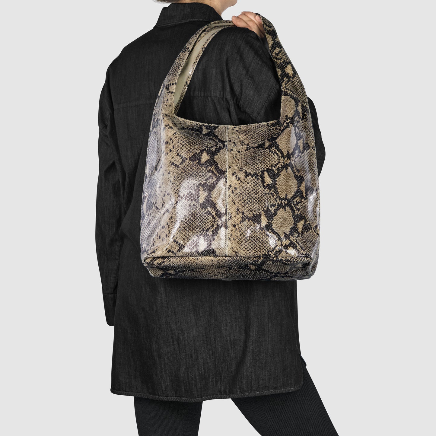 A person in black holds a statement shoulder bag with a brown and beige snakeskin pattern, likely a python embossed leather bag, slung over their shoulder, facing away from the camera against a plain light background.