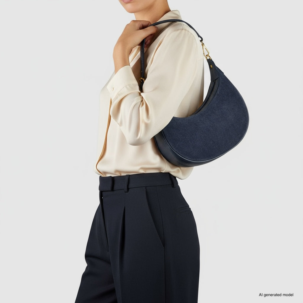 A woman in a cream blouse and dark trousers holds a suede crossbody bag, posing against a plain light background. Her face is partially out of frame.