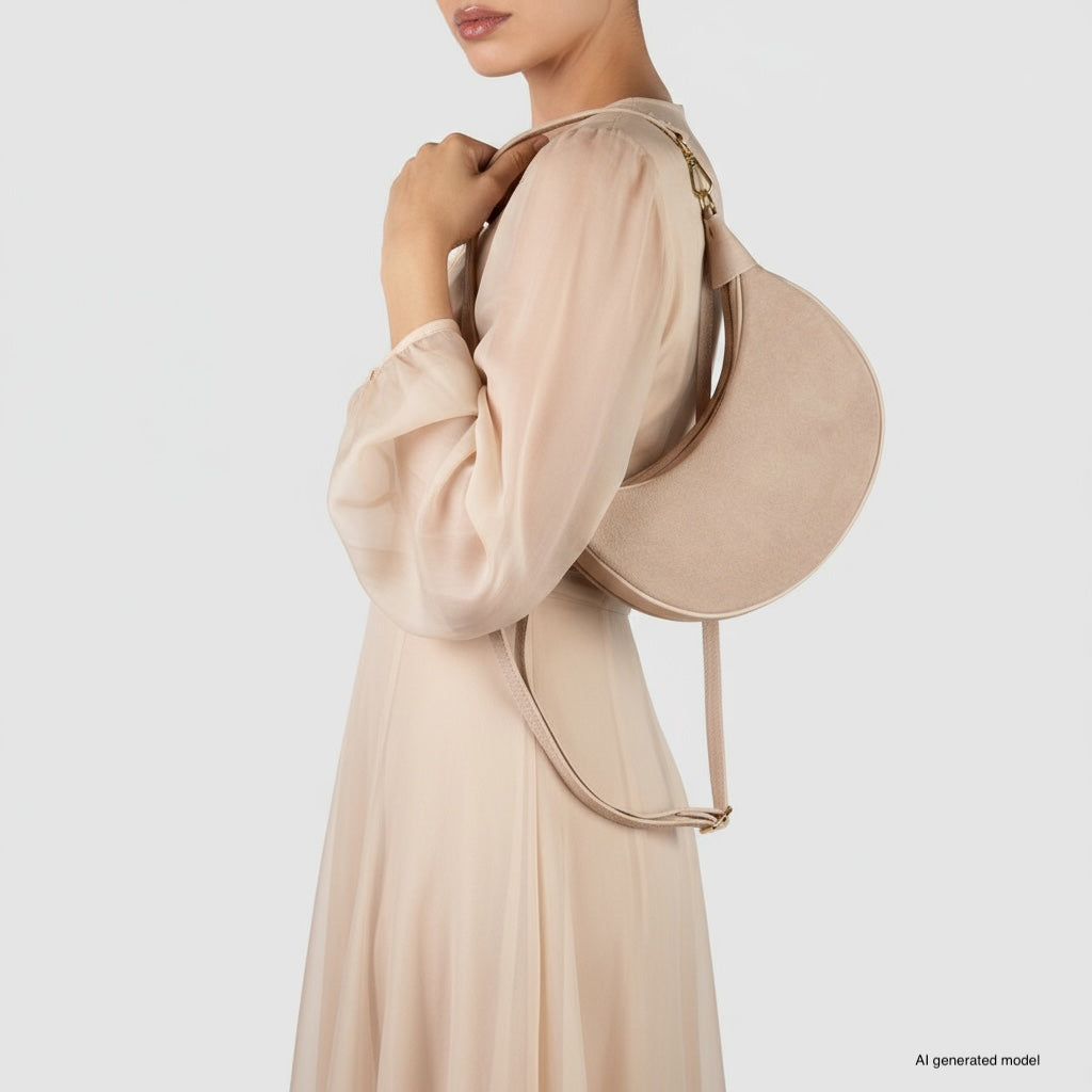 A woman in a long, sheer, beige dress holds a chic Italian leather crescent handbag over her shoulder, standing against a plain white background. Only her upper body and part of her face are visible.