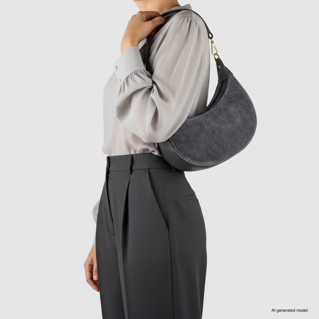 A person wearing a light gray blouse and high-waisted dark gray trousers holds an elegant Italian leather crescent handbag on their arm against a plain white background.