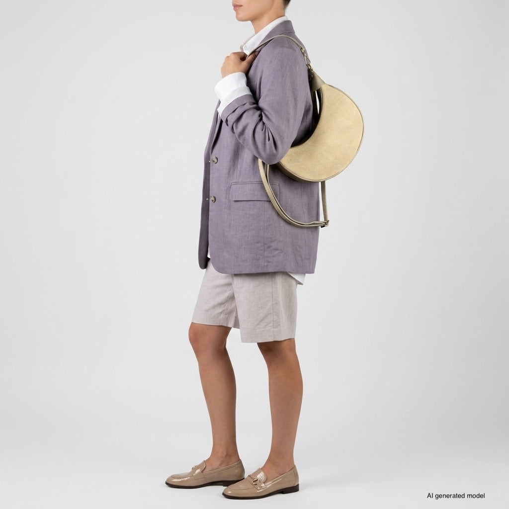A person wearing a light purple blazer, white shirt, beige shorts, and tan loafers stands against a plain background, carrying a large crescent handbag in beige.