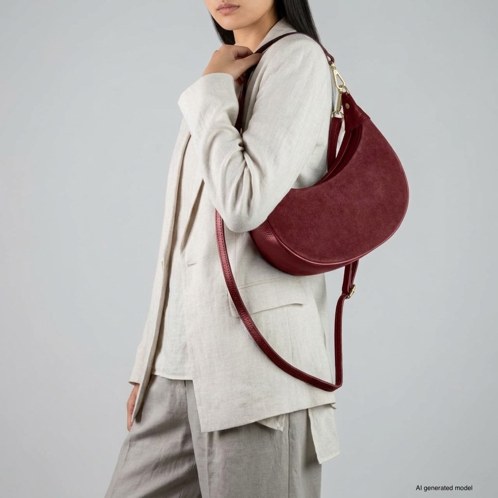 A woman in a light beige suit with loose trousers holds a crescent handbag crafted from Italian leather over her shoulder. The background is plain gray.